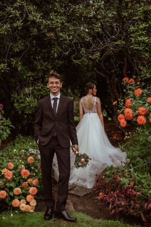 A couple in wedding attire walks through a lush garden path with vibrant orange dahlias blooming alongside them.