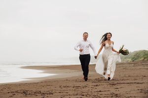 Two people in wedding attire run playfully along a scenic beach at sunset.