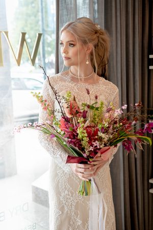 Bride in elegant lace gown holds vibrant burgundy and pink floral bouquet with cascading greenery against window light.