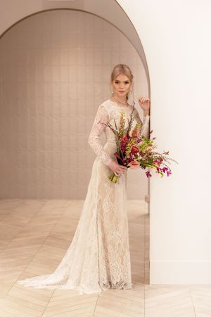 Elegant bride in cream lace gown with long sleeves holds colorful floral bouquet against neutral arched backdrop.