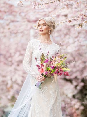 A bride in a lace wedding gown holds a vibrant spring bouquet beneath blooming cherry blossoms in a dreamy garden setting.