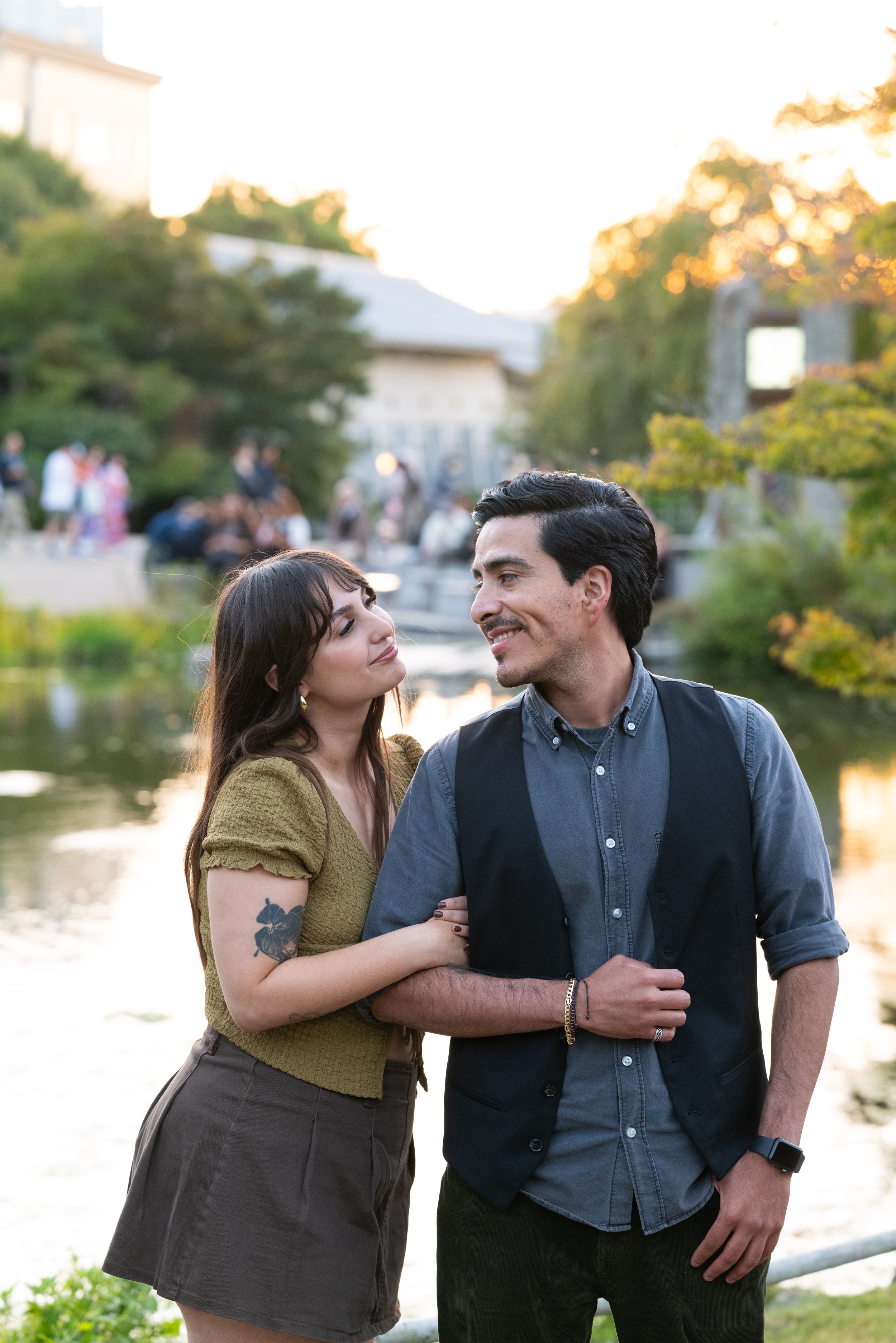 Couple sharing a loving glance while embracing near a reflective pond at golden hour in a park.