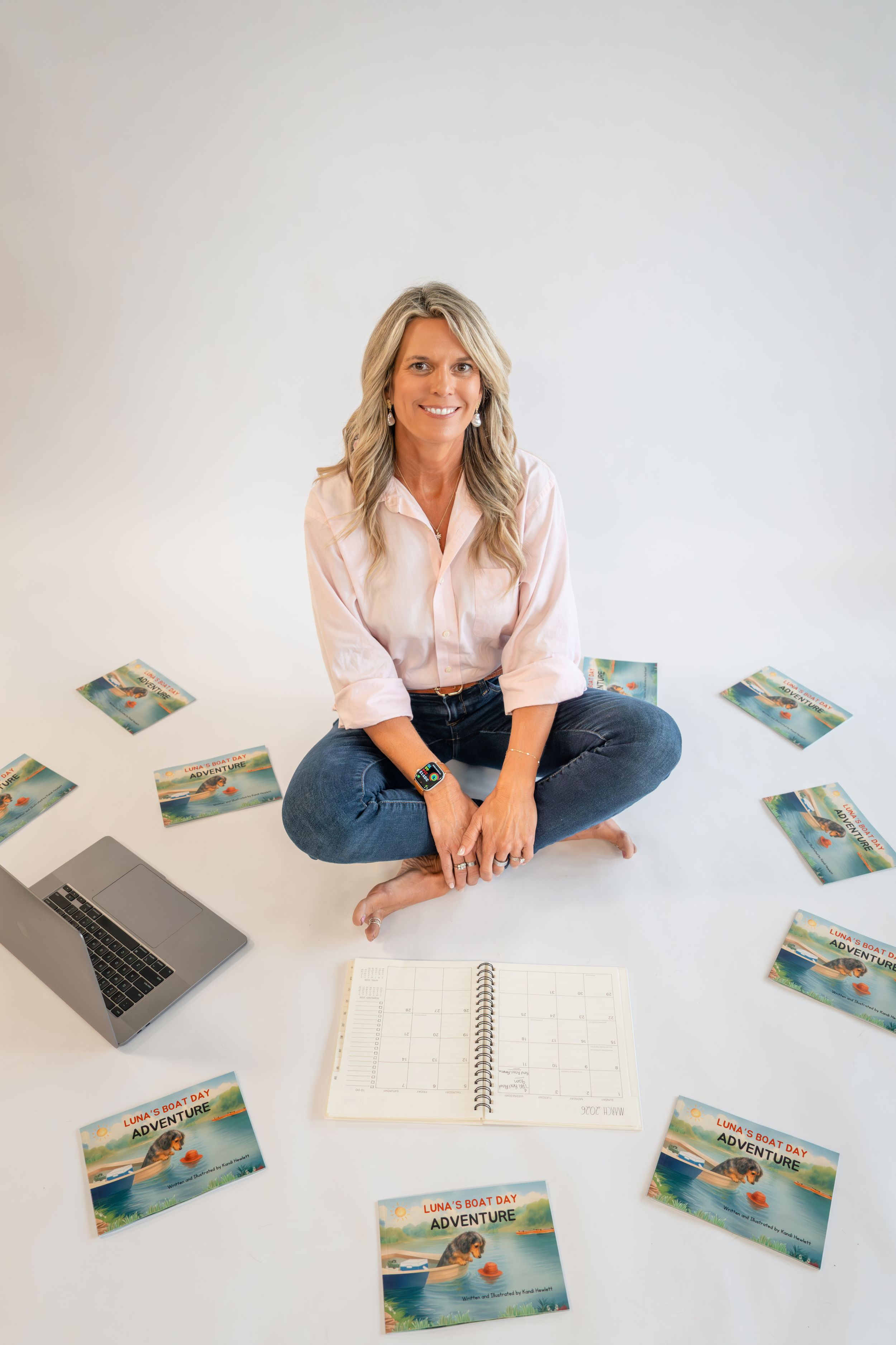 Woman sitting cross-legged on white floor surrounded by children's adventure books and a laptop, smiling at camera.