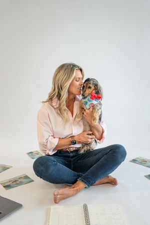 Woman sitting cross-legged on white floor kissing small dog wearing colorful bandana, surrounded by planners.