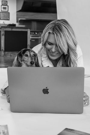 Black and white photo of smiling blonde woman with dachshund dog looking at Apple MacBook laptop at home.