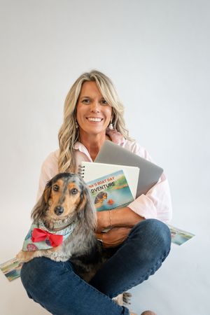 Smiling blonde woman sitting cross-legged holding a laptop and children's book, with a dachshund wearing a red bow tie.