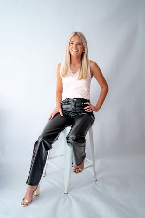Woman in pink tank top and leather pants sits confidently on white stool against light background.