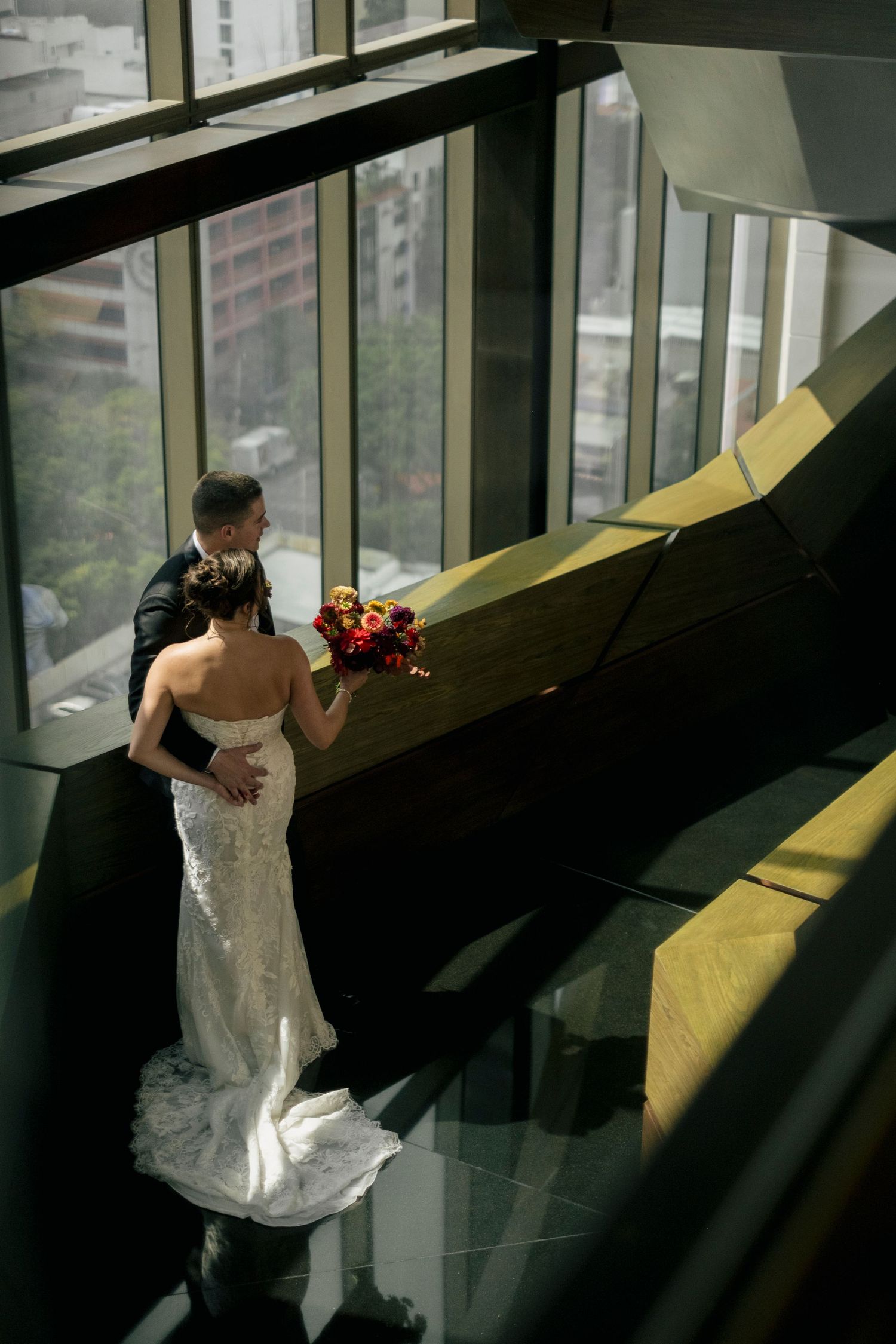 A bride in a white dress stands near large windows in a dramatic architectural space with angular shadows.