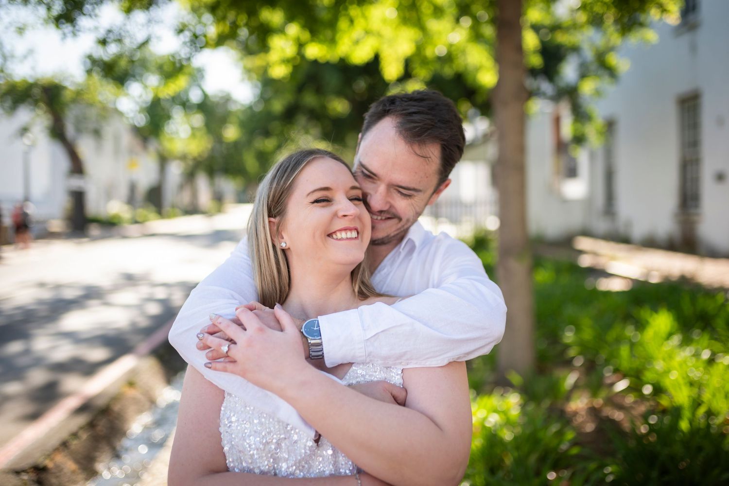 A couple embraces outdoors on a sunny day with lush greenery and white buildings in the background.