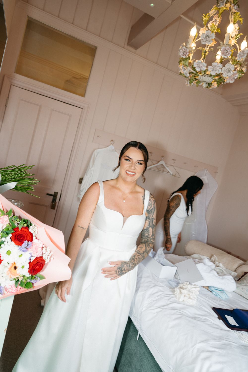 A wedding dress fitting scene with a white gown, floral bouquet and elegant chandelier in a bright interior room.
