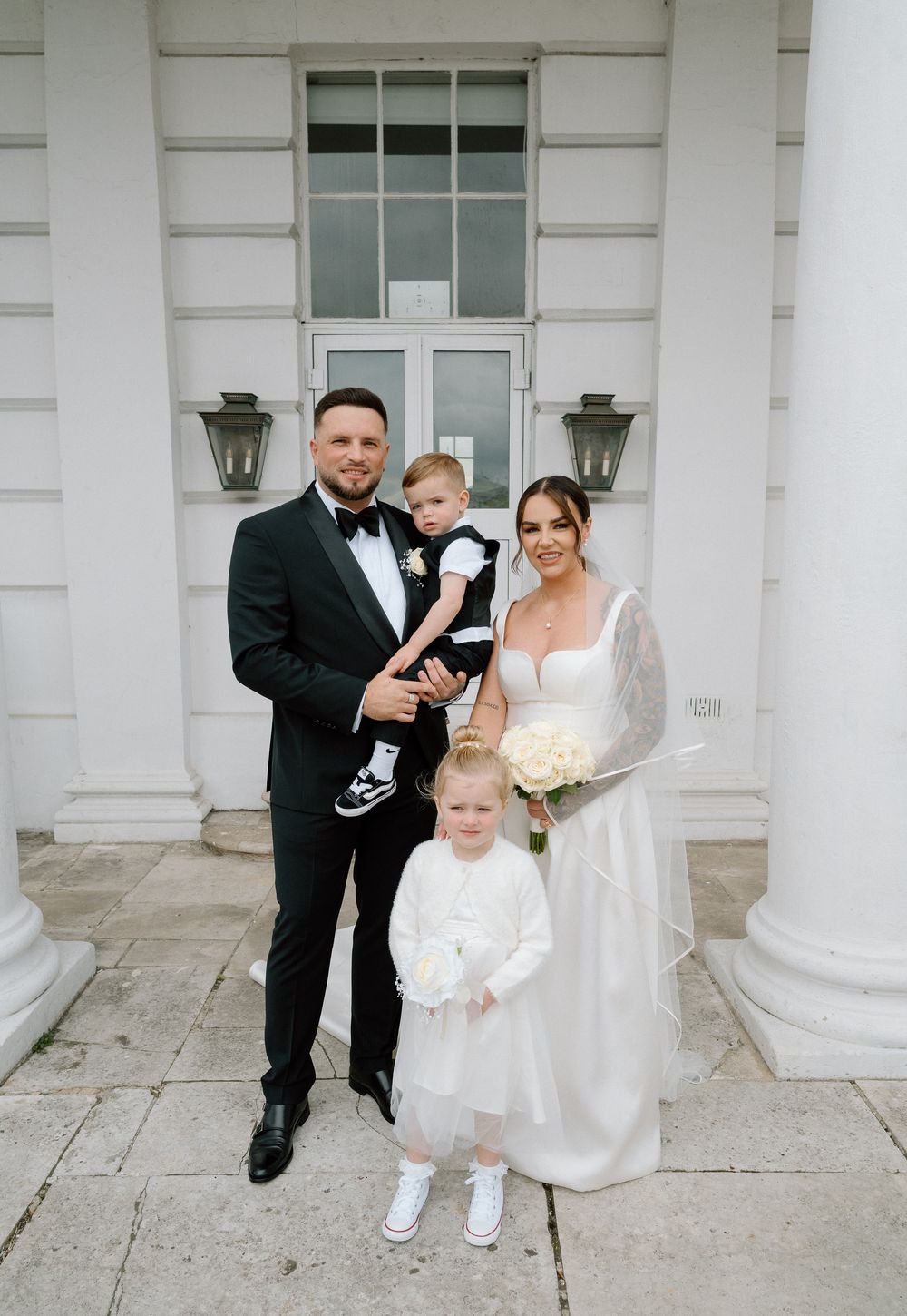 Elegant wedding portrait of a family in formal attire standing in front of a white columned entrance with lanterns.