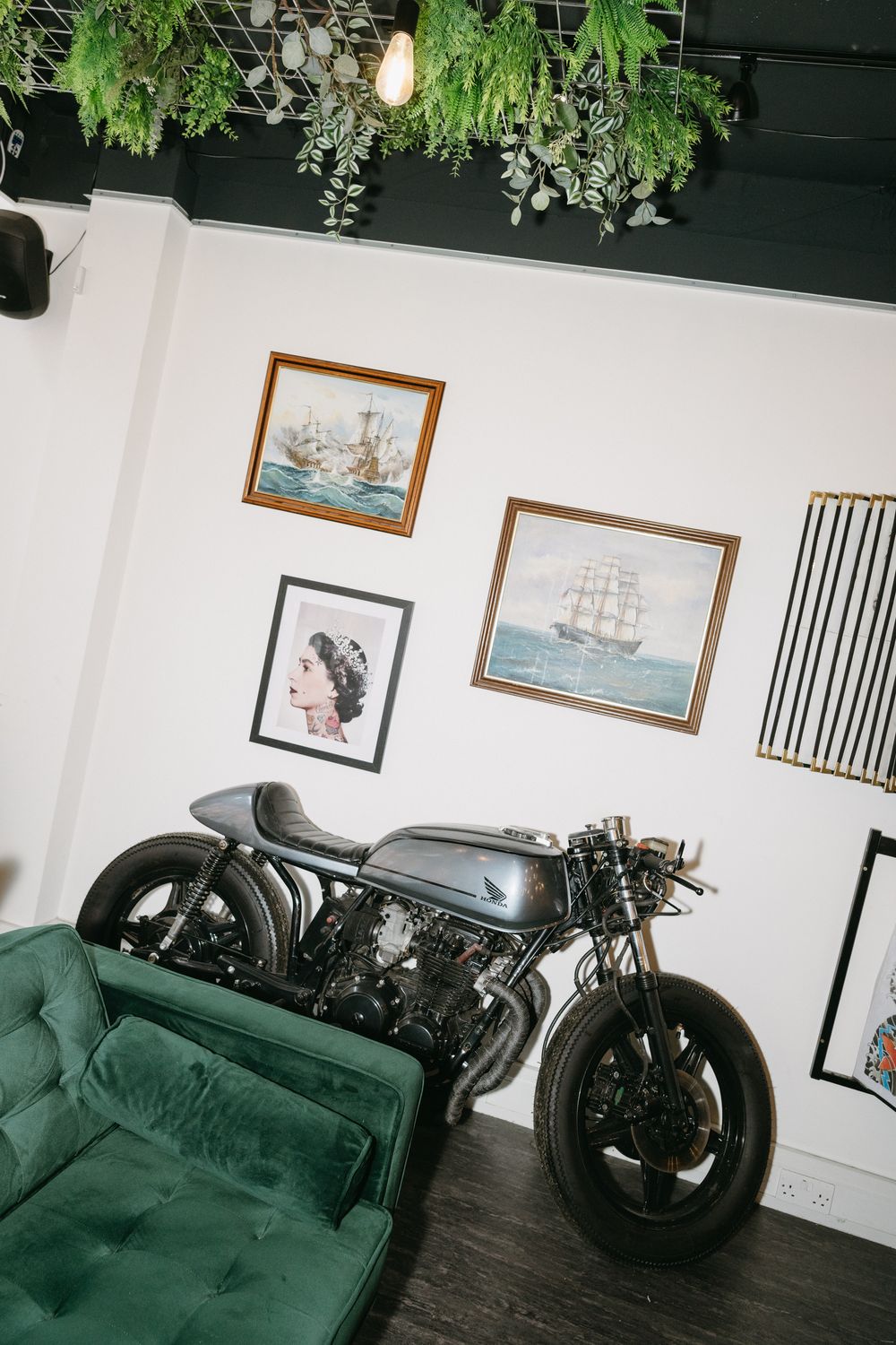 Vintage motorcycle parked next to green velvet couch with nautical artwork and hanging plants decorating white walls.