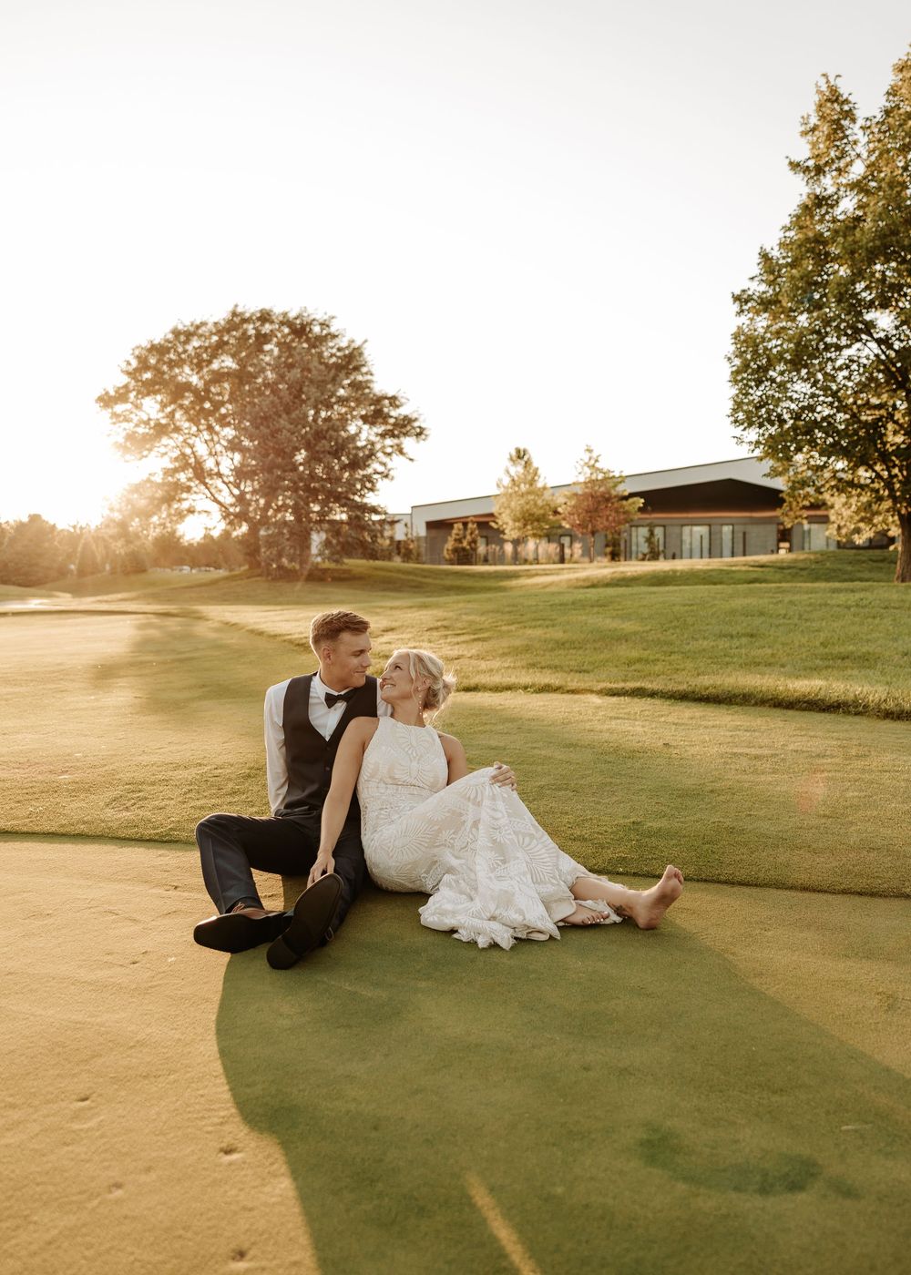 A couple sits together on a golf course at sunset with a clubhouse visible in the background.