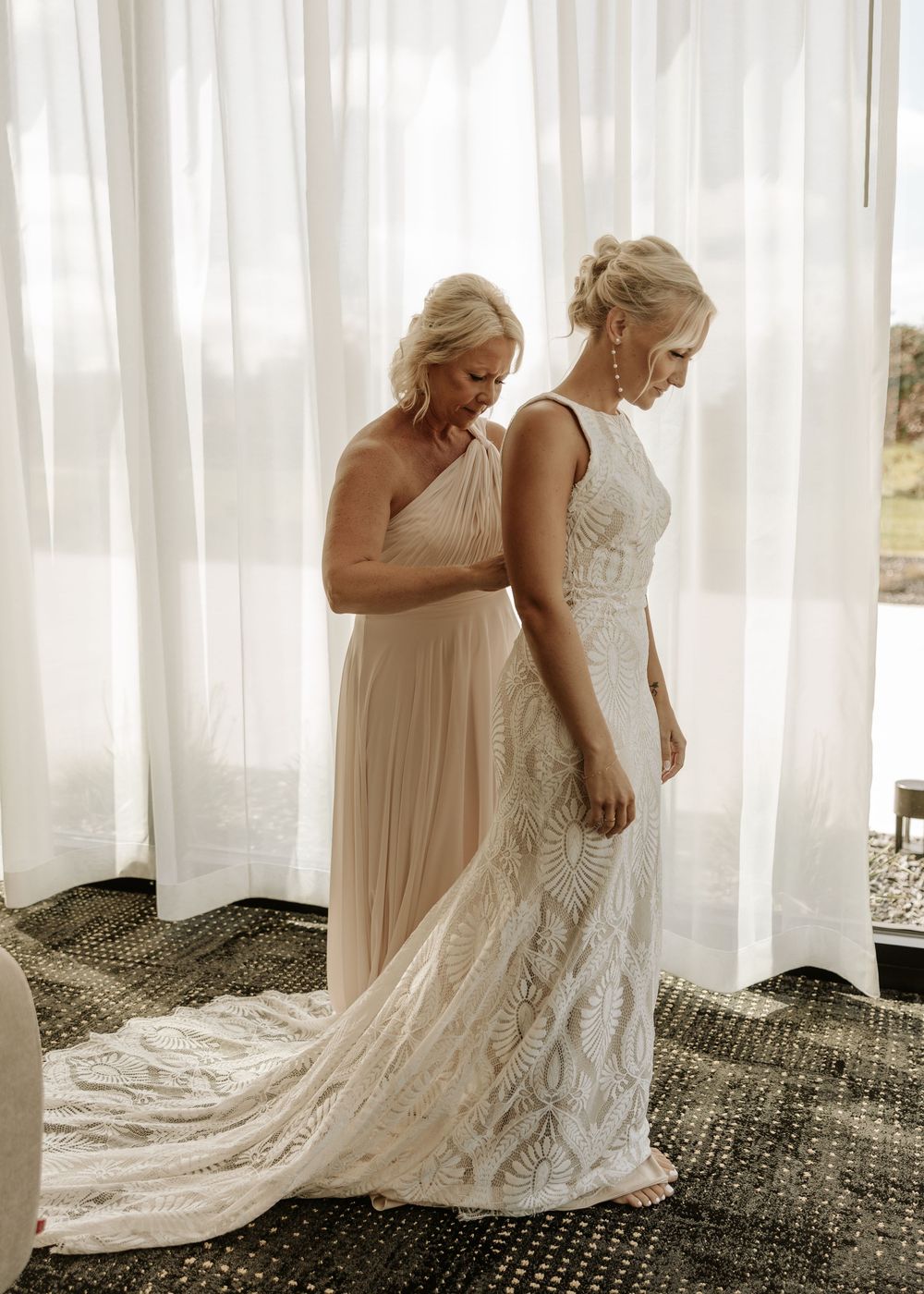 Two women in formal dresses prepare for a wedding against white curtains.