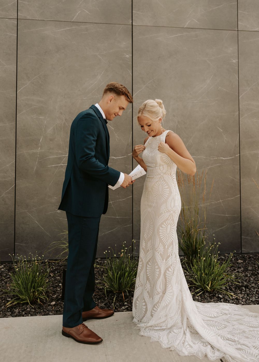 A wedding couple shares an emotional first look moment against a grey wall with greenery.