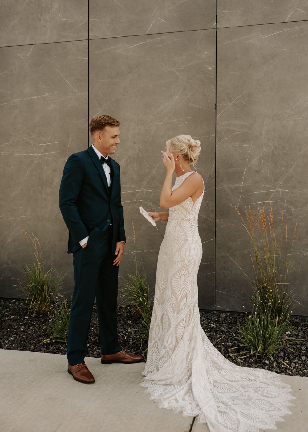 Wedding couple interacts playfully against grey stone wall with landscaping.