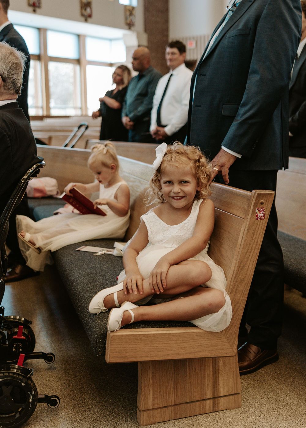 A young flower girl in a white dress sits playfully on a wooden church pew during a wedding ceremony.