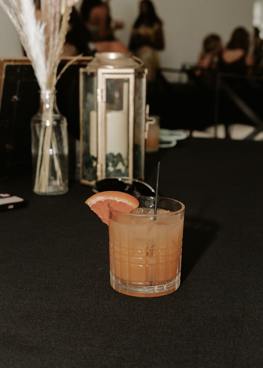 Pink cocktail with grapefruit garnish sits on black table with decorative lantern and dried flowers.