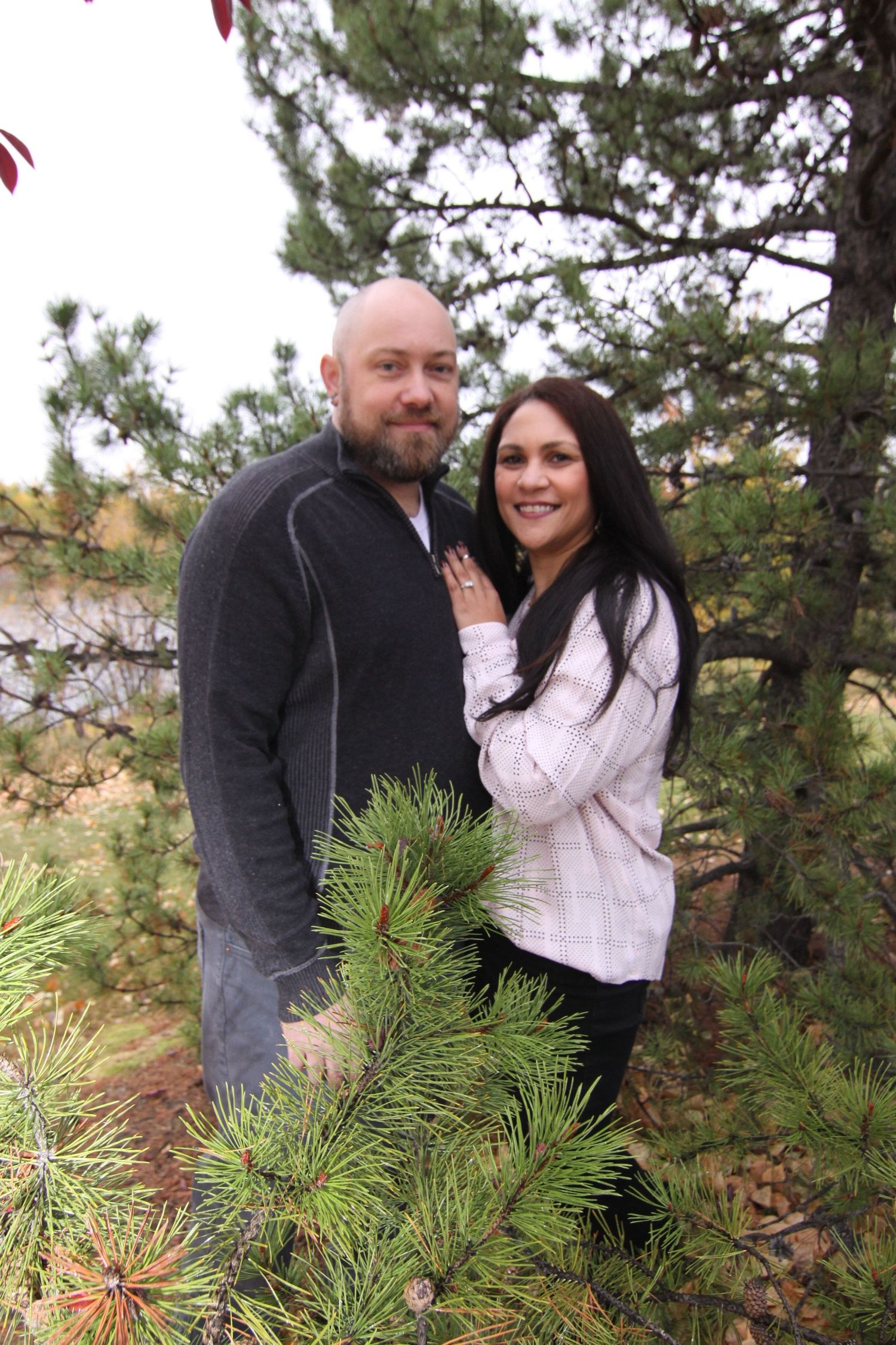 A couple poses together outdoors among evergreen pine trees during a winter photo session.