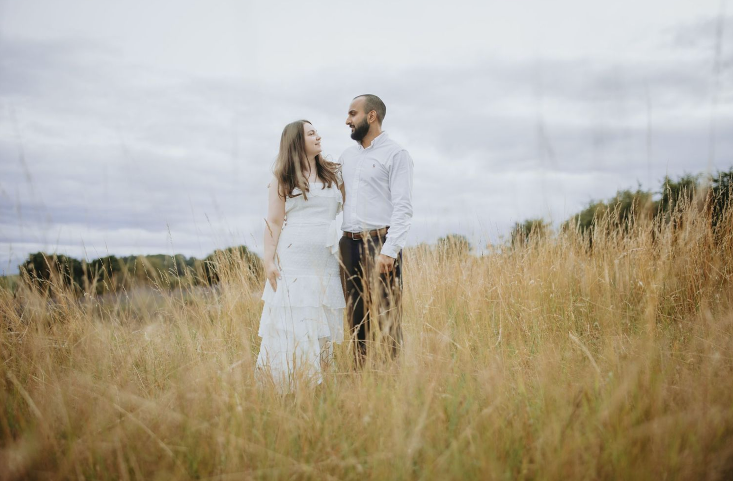 Couple walks through tall golden grass in a meadow during sunset, creating a romantic silhouette against the sky.