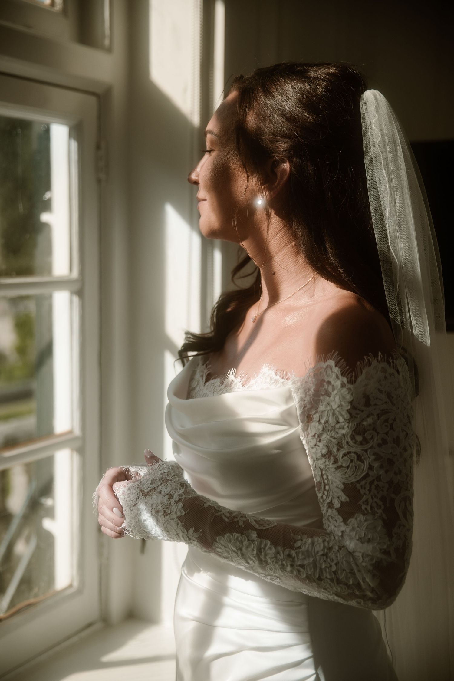 A bride in a white lace-sleeved wedding dress gazes thoughtfully out a sunlit window in a romantic portrait.