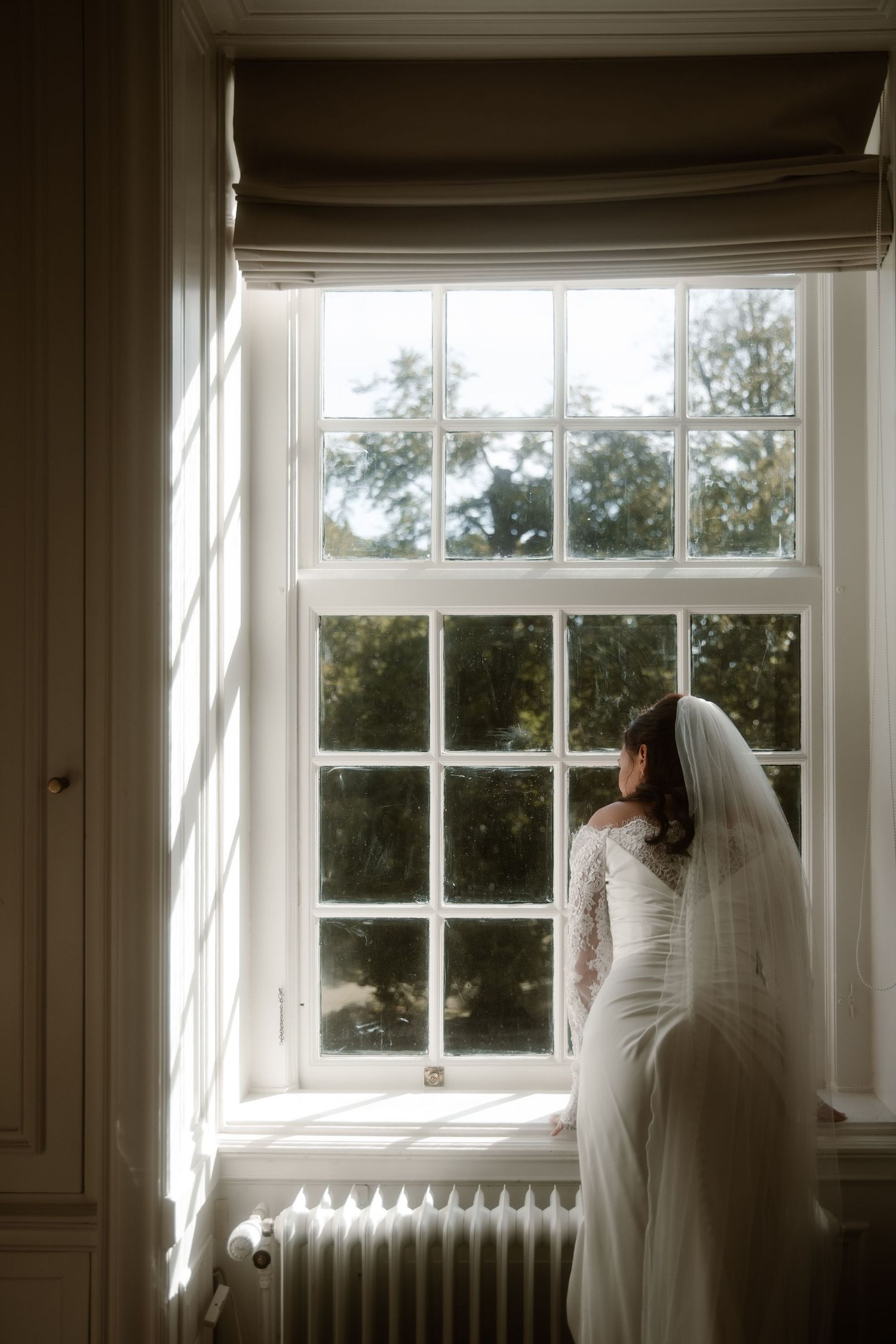A ghostly figure in white stands by a bright window in a minimalist interior space.