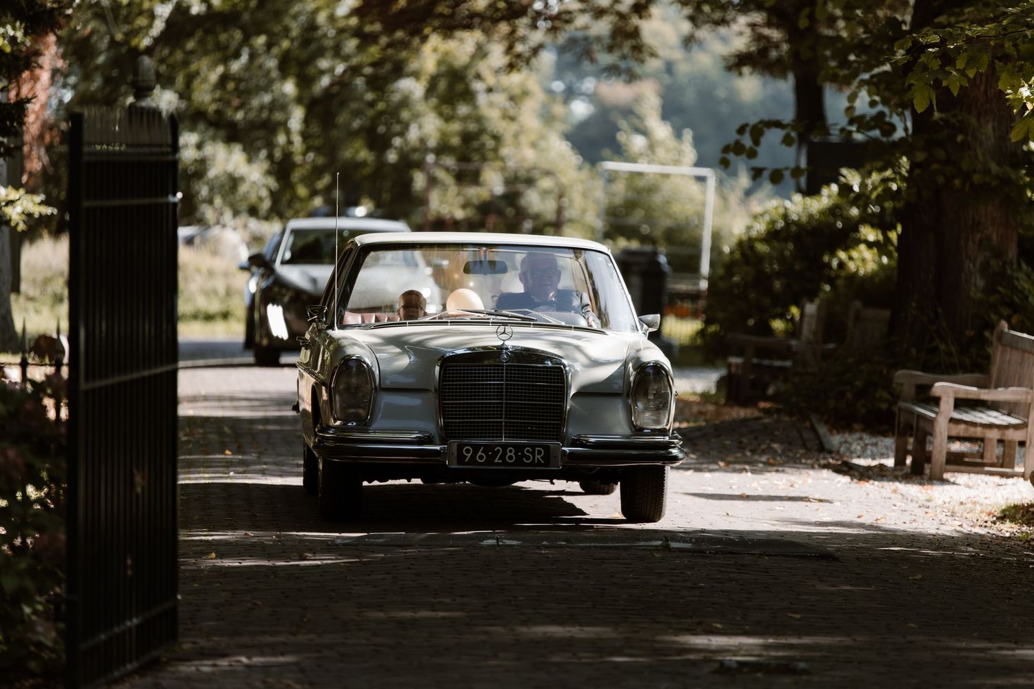 A classic vintage Rolls-Royce car parked in a shaded tree-lined driveway on a sunny day.
