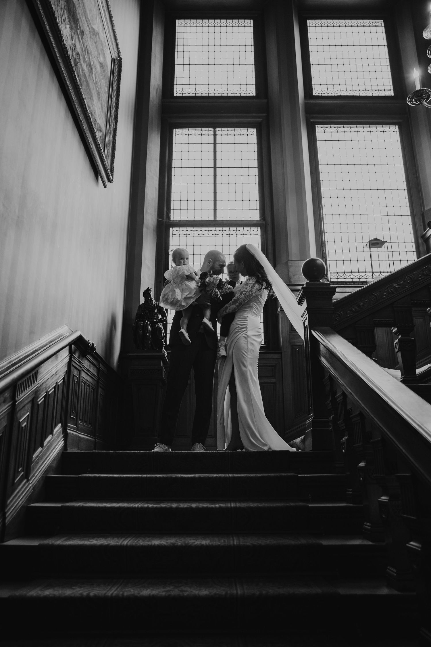 Silhouettes of wedding guests gather by large windows in a dramatic black and white composition.