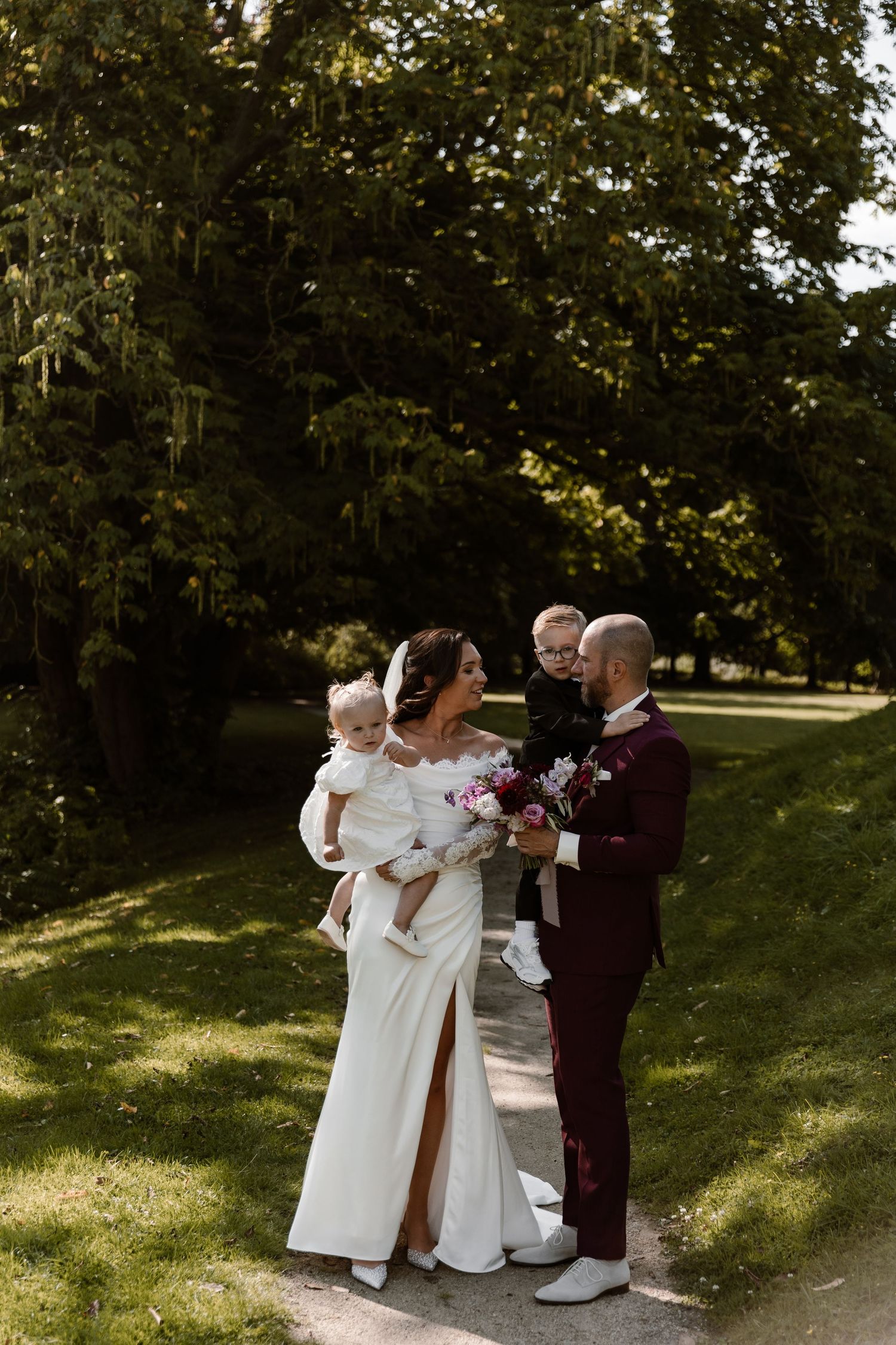 Wedding party members in white and burgundy attire pose together on a sunny path surrounded by trees.