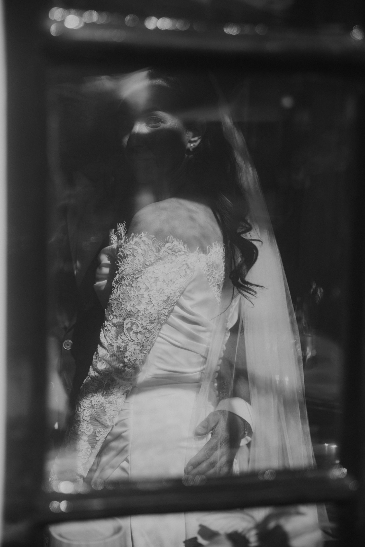 Black and white artistic wedding photo showing a bride in white dress through a car window reflection.