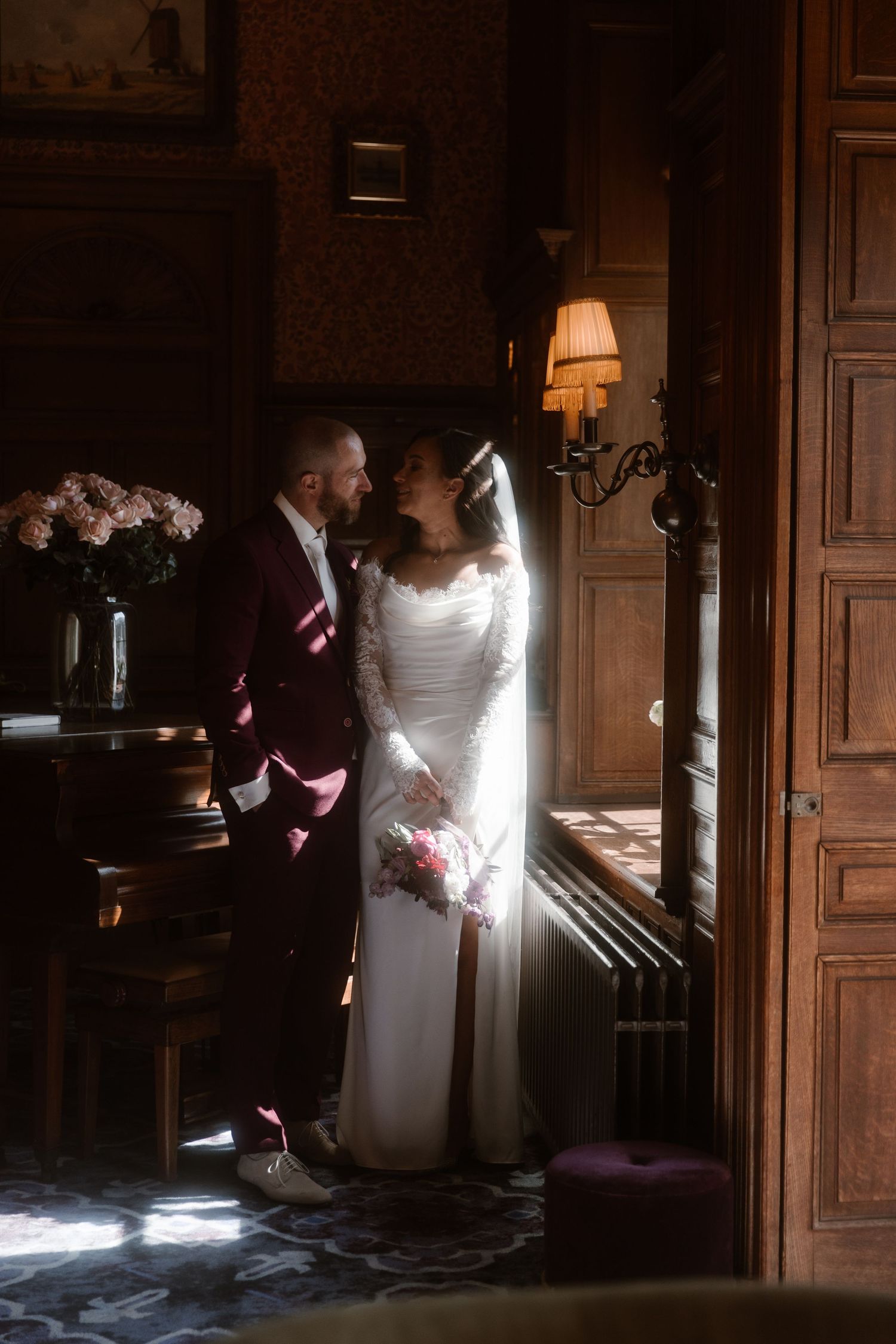 Sunlight streams through windows illuminating a bride in a white dress standing in an elegant dark wood room.