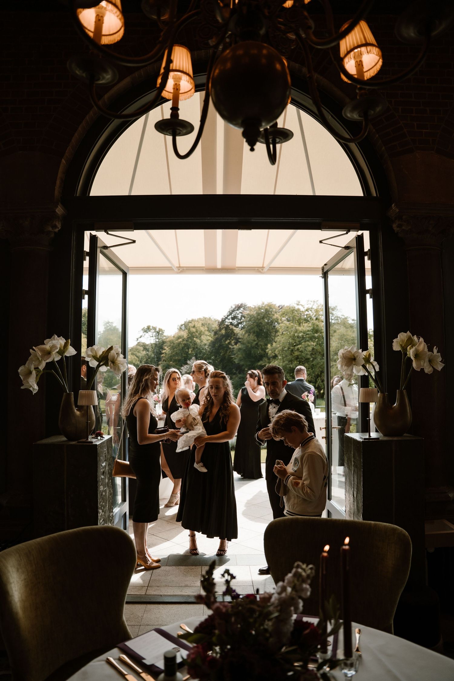 Silhouettes of guests gathered in a dark reception venue with bright doorways and windows creating dramatic lighting.