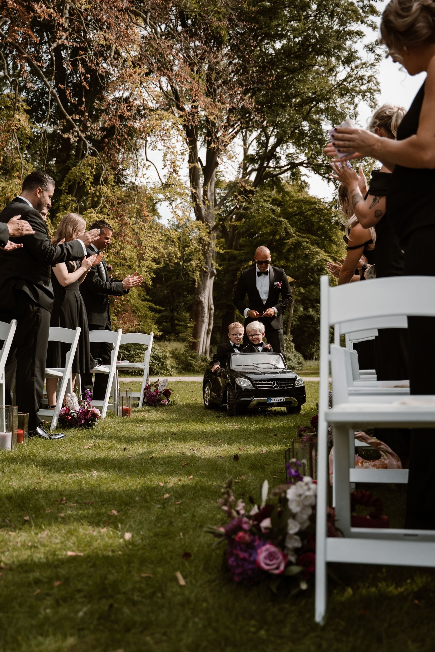 A wedding ceremony takes place outdoors with white chairs arranged on a grassy lawn surrounded by trees.