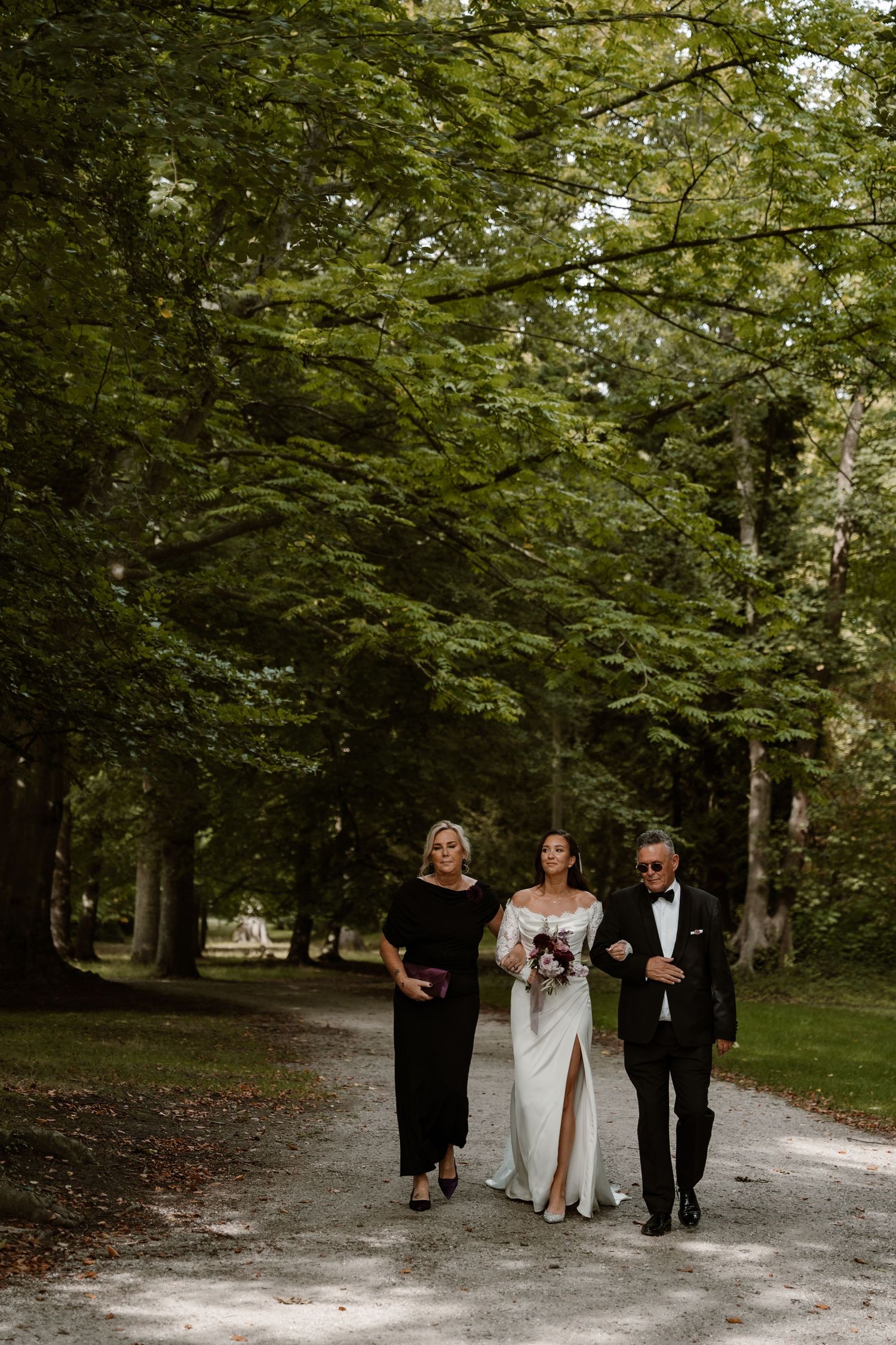 Three elegantly dressed figures walk together down a tree-lined path during a formal outdoor event.