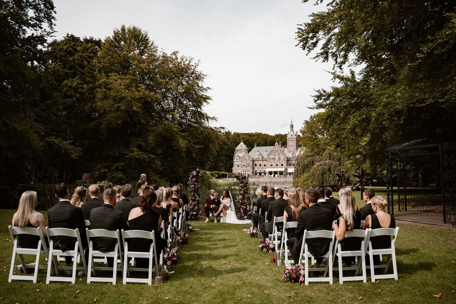 Outdoor wedding ceremony setup with white chairs arranged in rows on a grassy lawn surrounded by tall trees.
