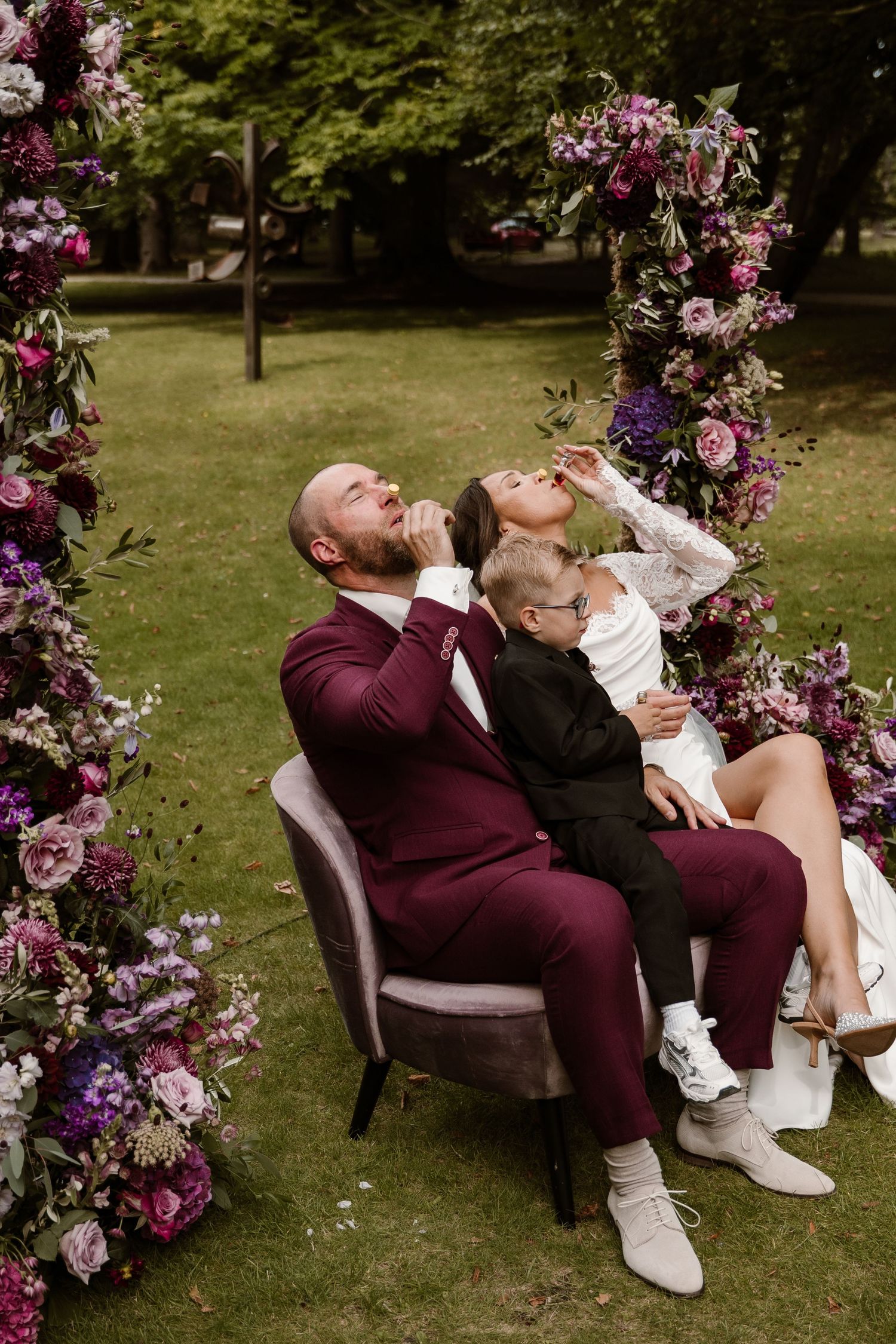 A romantic wedding couple shares intimate moments on vintage chairs surrounded by pink floral archways in a garden setting.