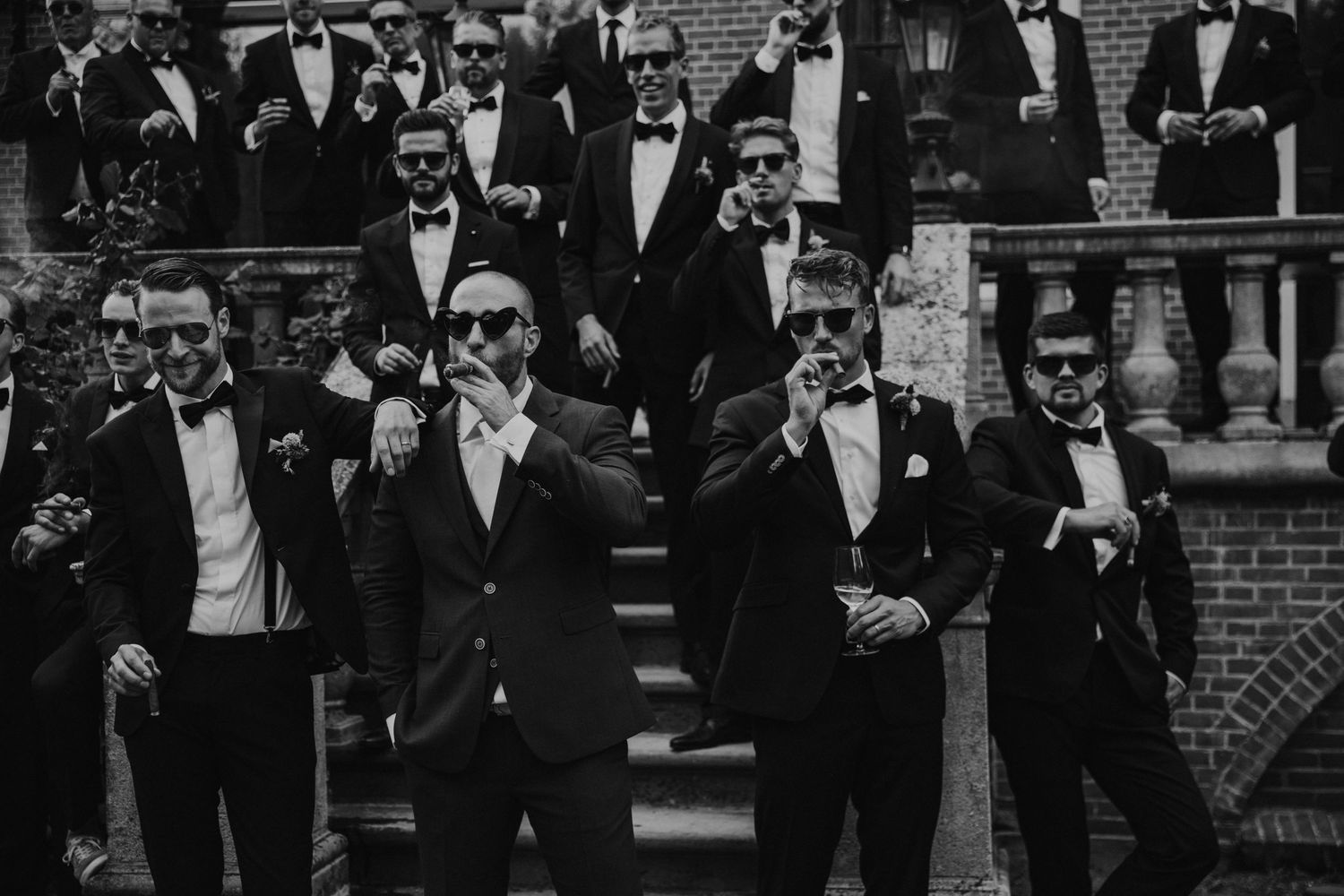 Groomsmen in black tuxedos and sunglasses pose on brick steps in a stylish black and white photo.