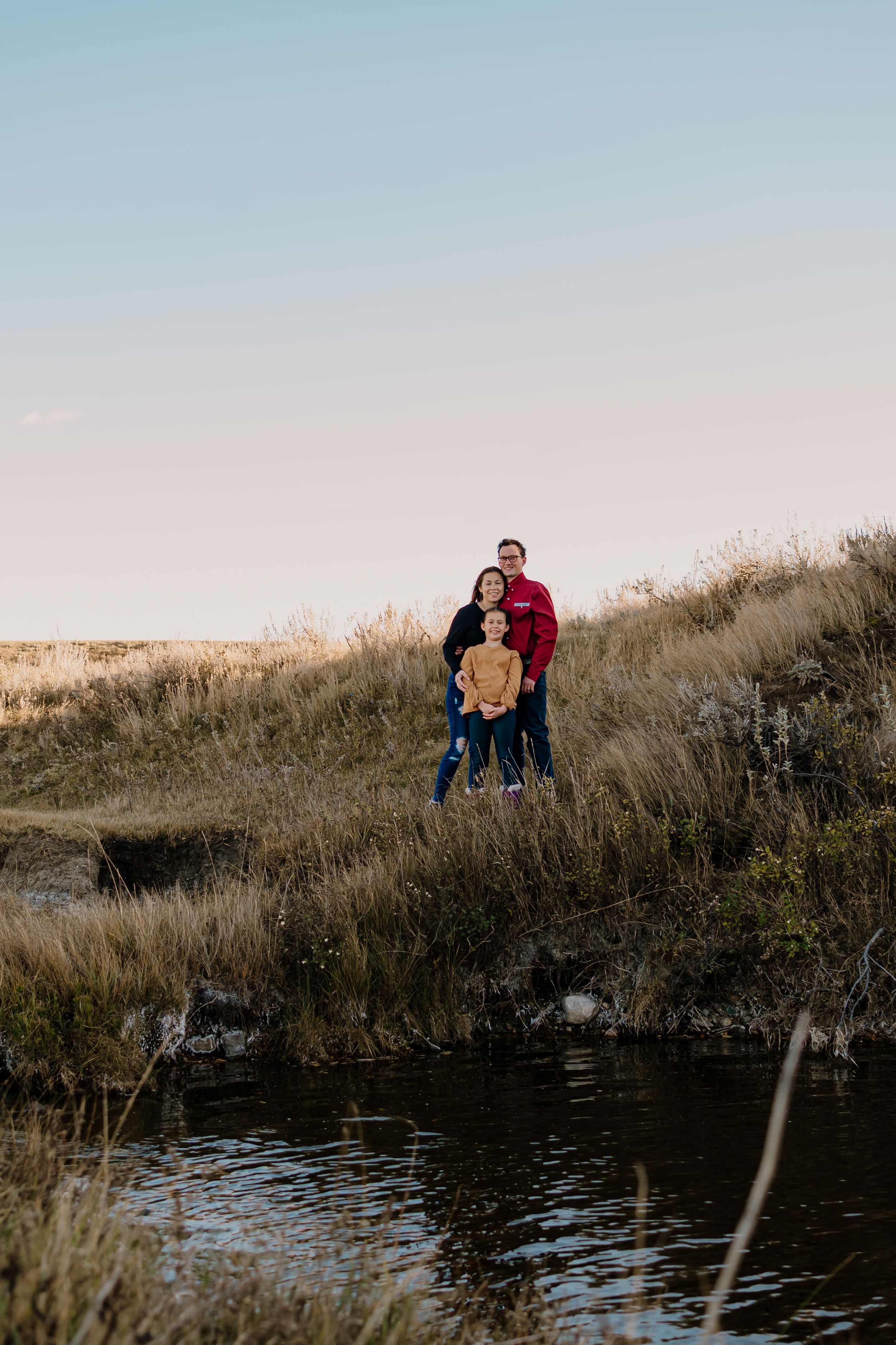 A family walks together along a grassy hillside near a dark pond during golden hour.