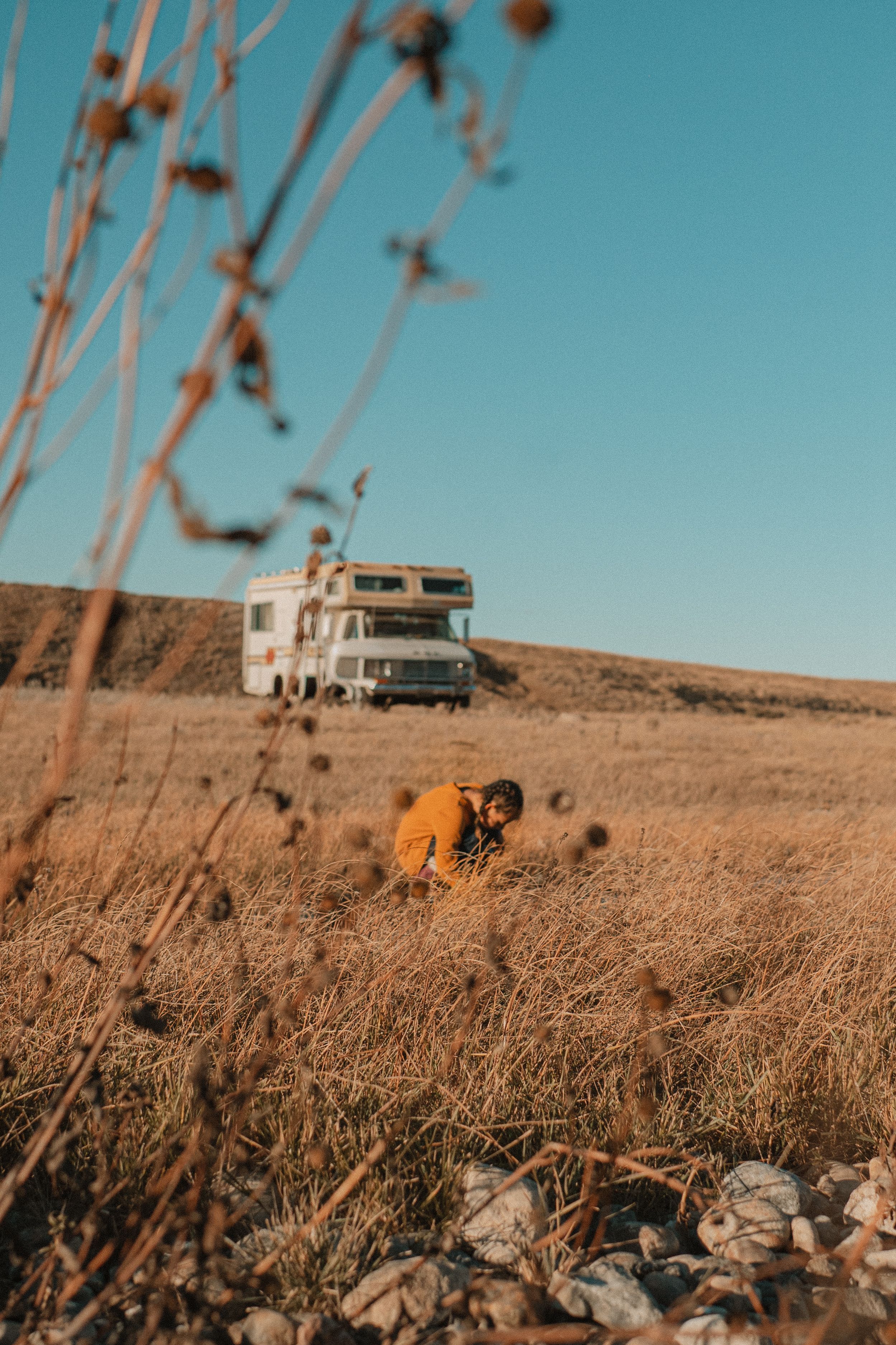 A vintage camper van sits on a hilltop against a bright turquoise sky with dried grass in the foreground.