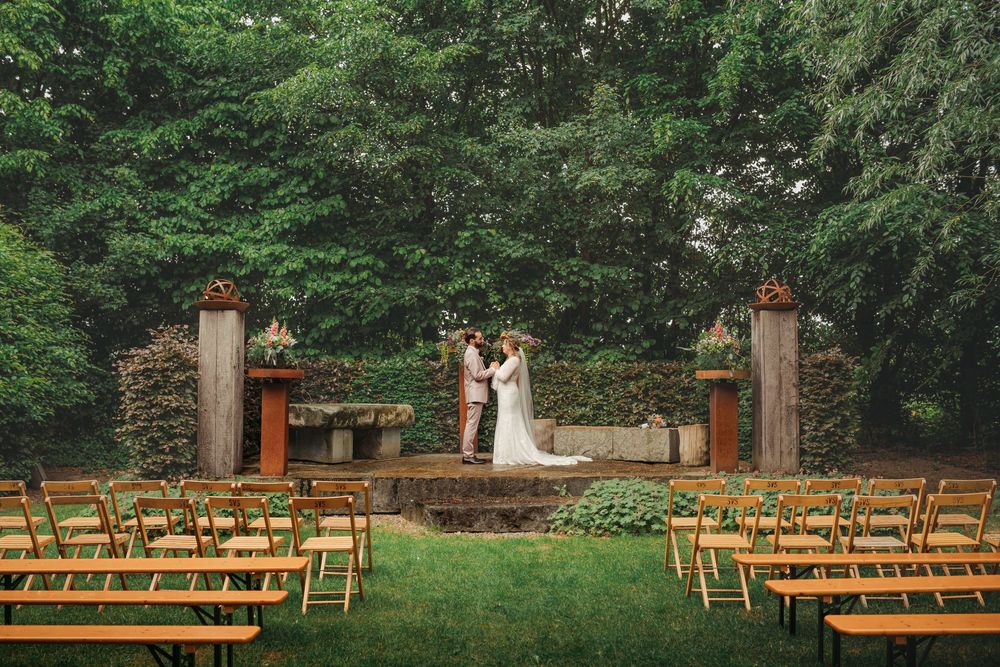 Wooden chairs arranged in rows for an outdoor wedding ceremony in a lush garden setting with stone pillars and greenery.