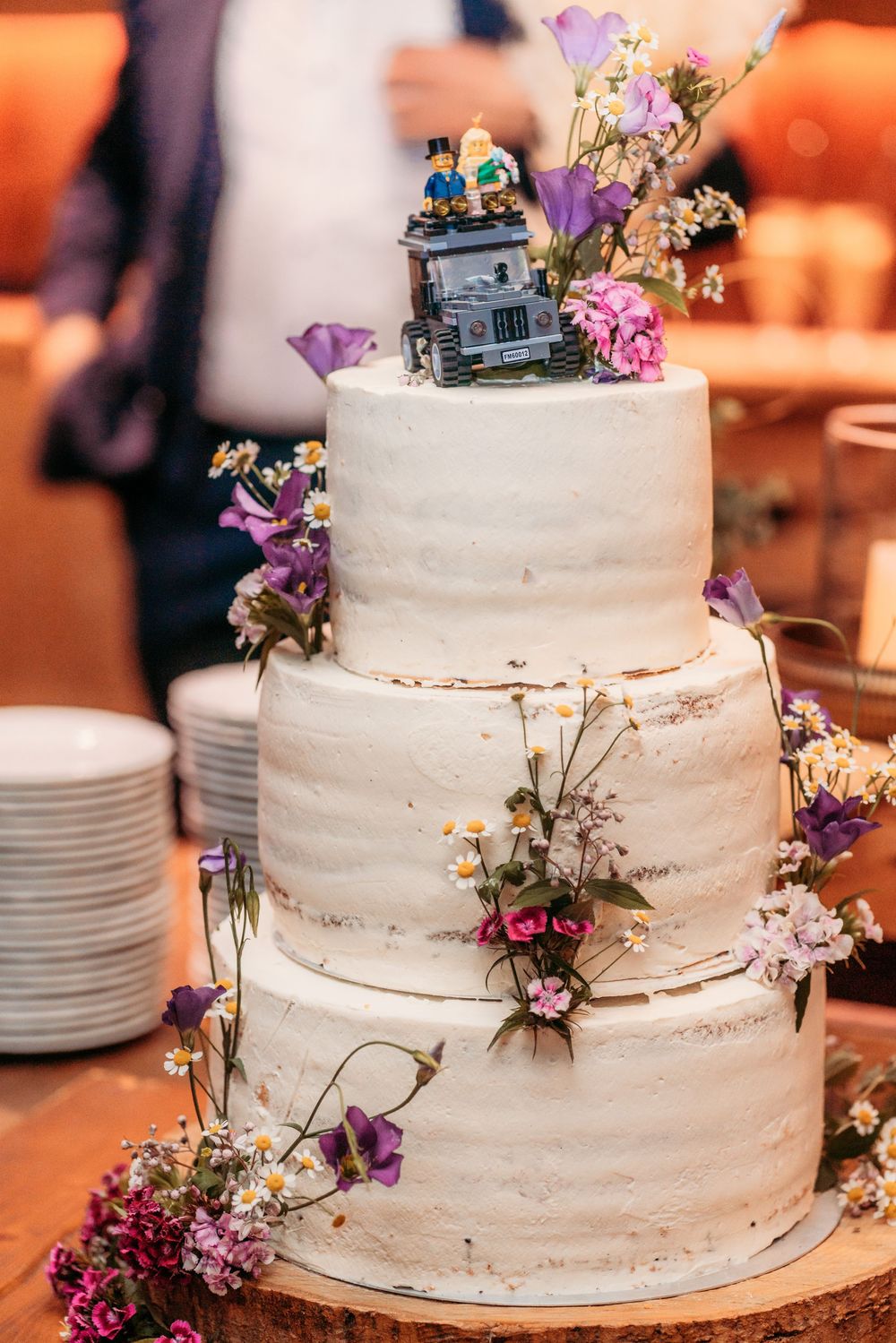 A white tiered wedding cake decorated with purple and pink wildflowers and delicate floral accents on a wooden cake stand.