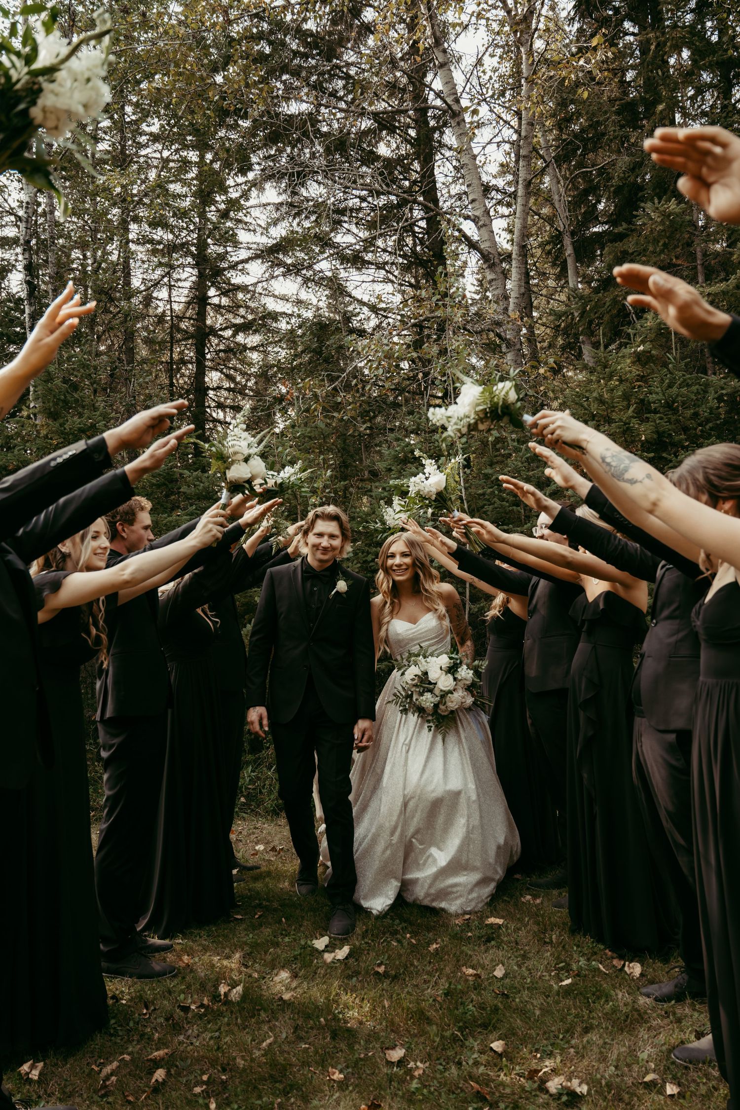 Wedding party in dark attire tossing flower petals over a couple walking through a wooded outdoor setting.