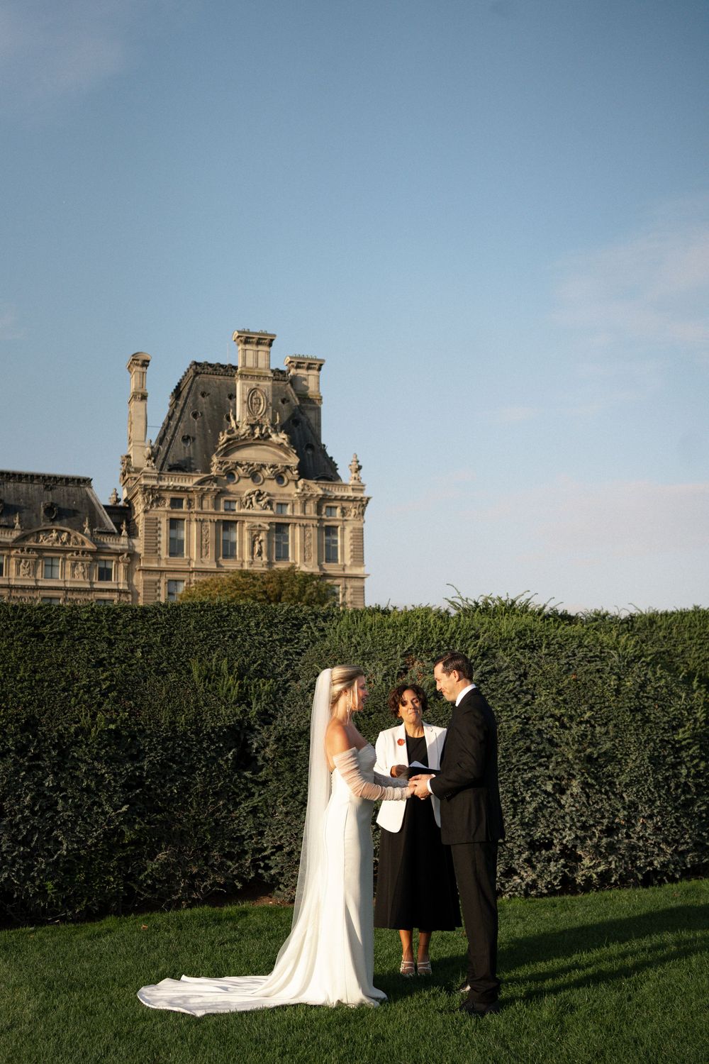 A wedding ceremony takes place on a manicured lawn with elegant French architecture visible in the background.Paris editorial elopement phot