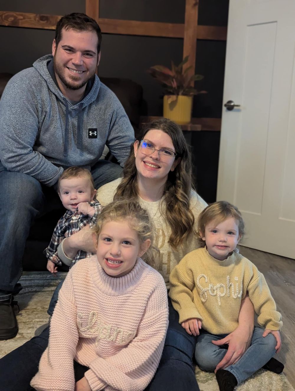 Family group sitting together on floor in casual indoor setting wearing coordinated sweaters.