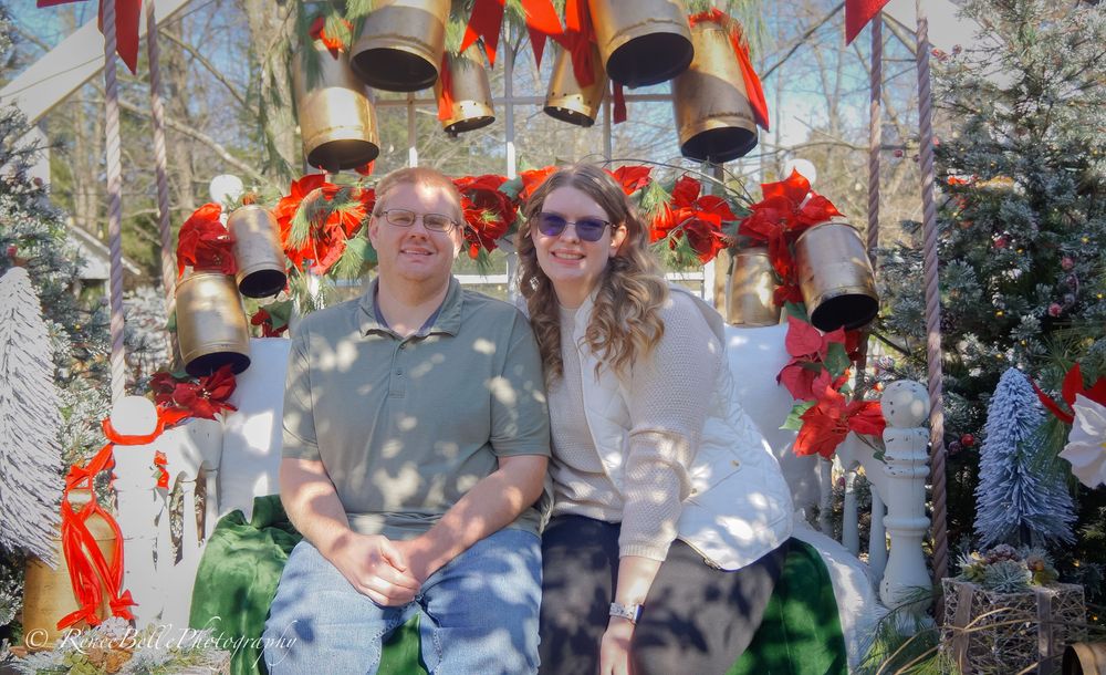 Two people sit beneath a festive arch decorated with red poinsettias and holiday bells in an outdoor garden setting.