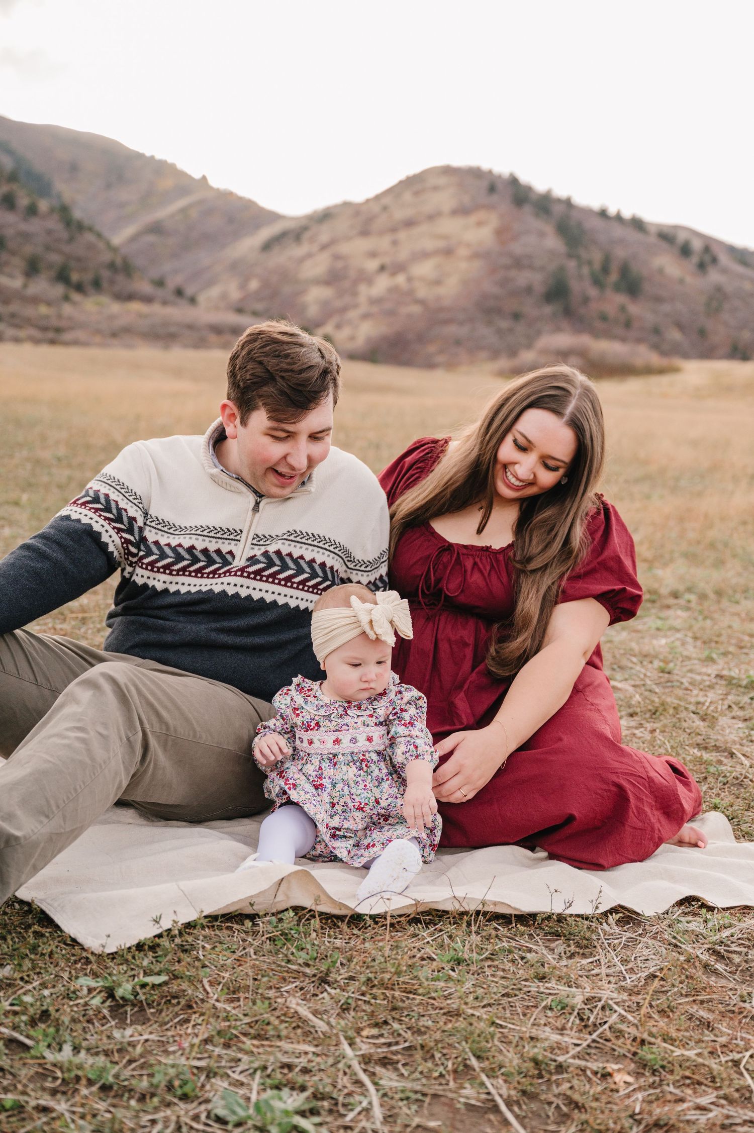 A family in coordinating winter sweaters relaxes together on a grassy hillside during an outdoor photo session.