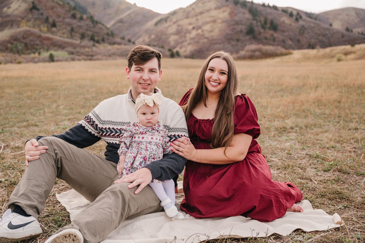 A family sits together on a blanket in a grassy field with mountains in the background during autumn.