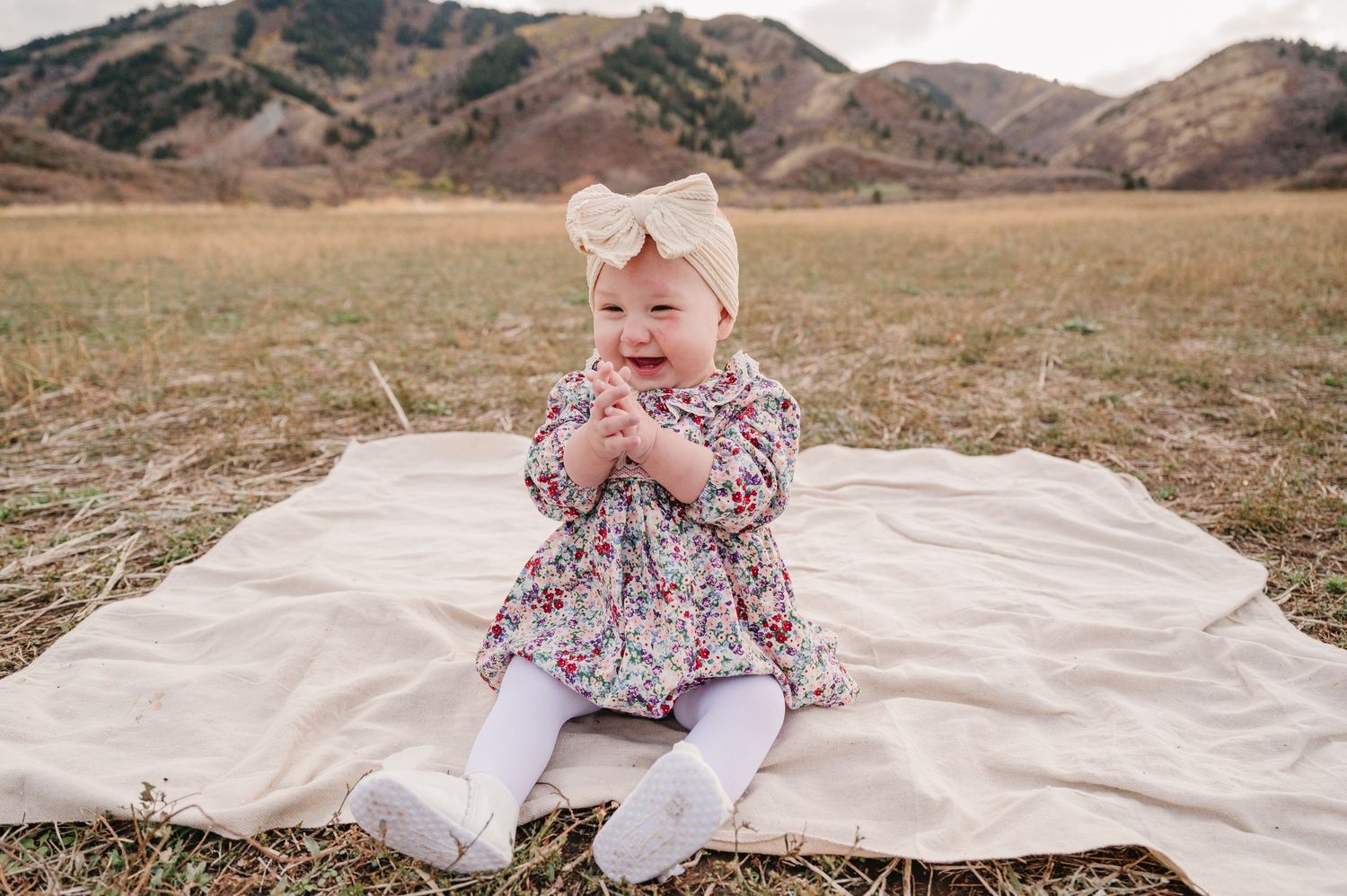 A family plays together on a white blanket in a grassy field with mountains visible in the distance.