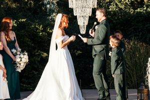Bride and groom exchanging vows outdoors at elegant wedding ceremony with chandelier, bridal party, and ring bearer present.
