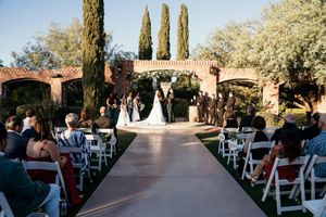 Outdoor wedding ceremony with guests seated in white chairs along an aisle leading to couple at floral arch.